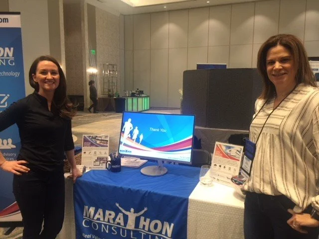 Two women standing at a booth with a blue tablecloth that reads "Marathon Consulting" at a conference.