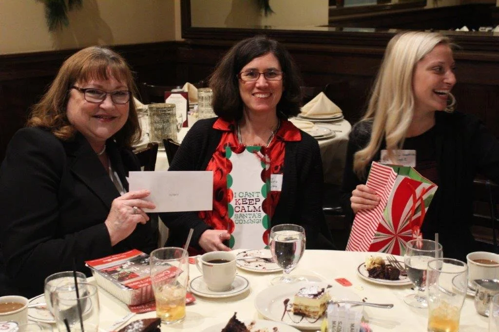 Three women sitting at a table during a holiday celebration, with Christmas-themed gifts, desserts, coffee, and drinks.