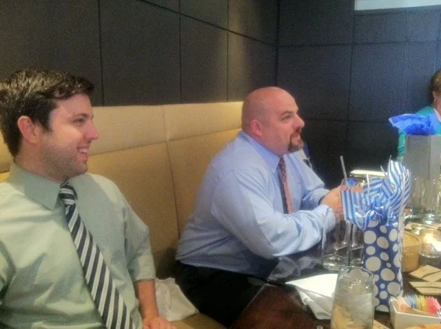 Two men sit at a table during a celebration, with wrapped gifts and decorations in the background.