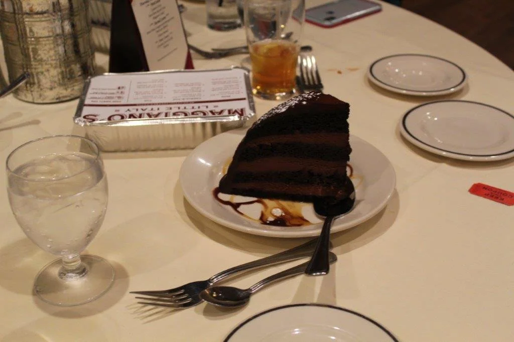 A slice of chocolate cake on a white plate with a spoon and fork, a glass of water, a glass with a beverage, and empty plates on a dining table.