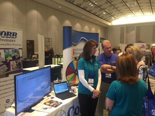 Group of people at a technology conference booth, engaging in conversation. The booth displays a spinning wheel and a monitor showing a software interface.