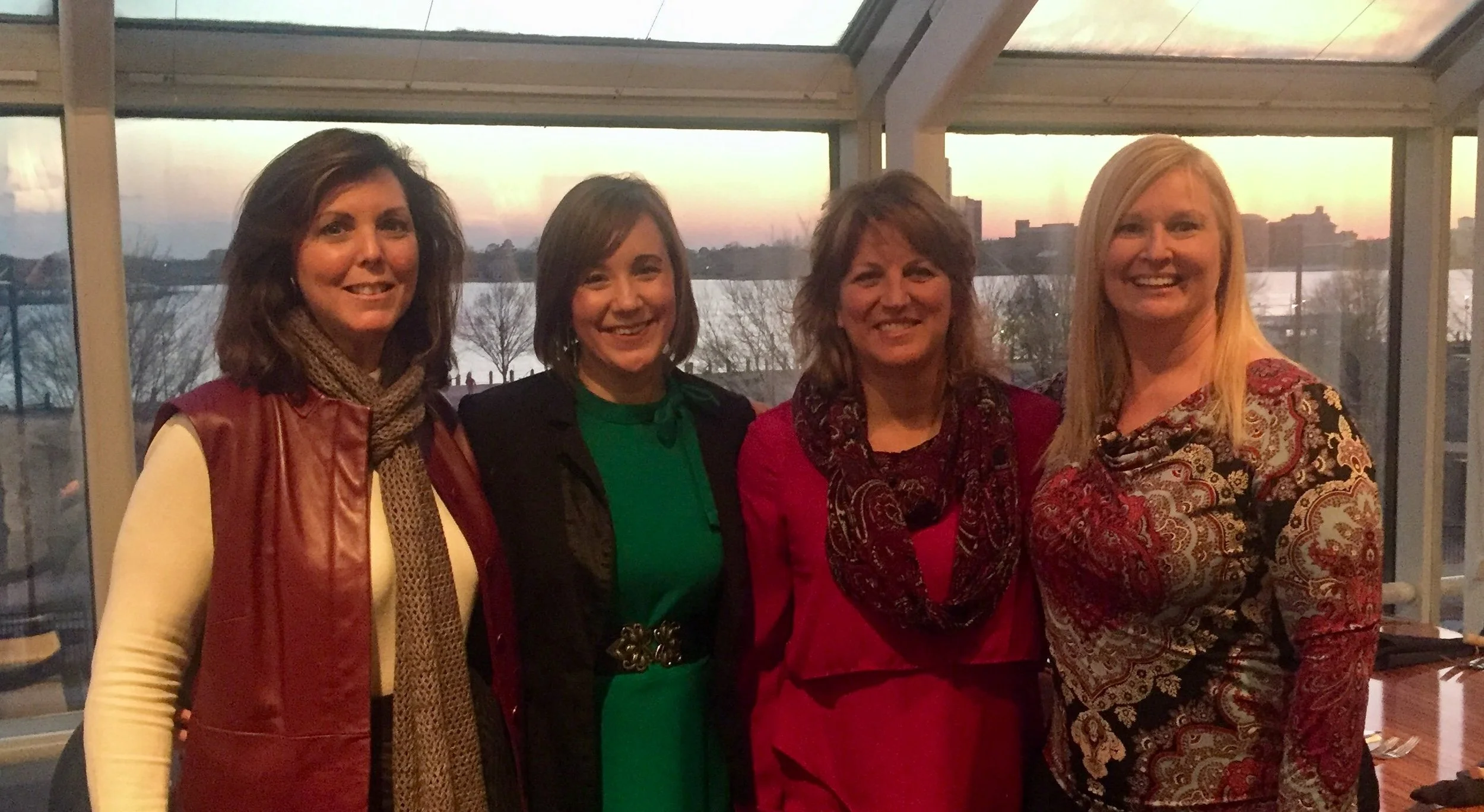 Group of four women standing together indoors with large windows and a sunset view of a river and trees in the background.