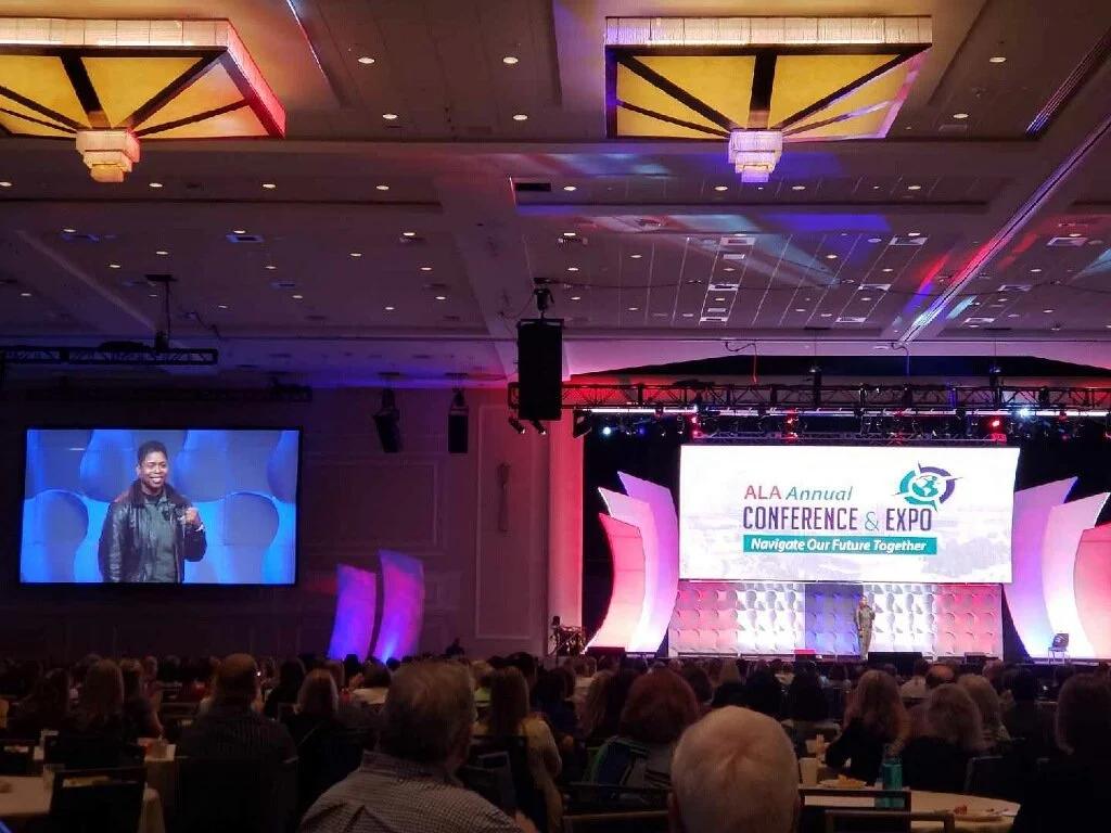 Large conference hall with many attendees, a stage with a big screen displaying 'ALA Annual Conference & Expo,' and a person speaking on stage with a smaller screen showing their live image.
