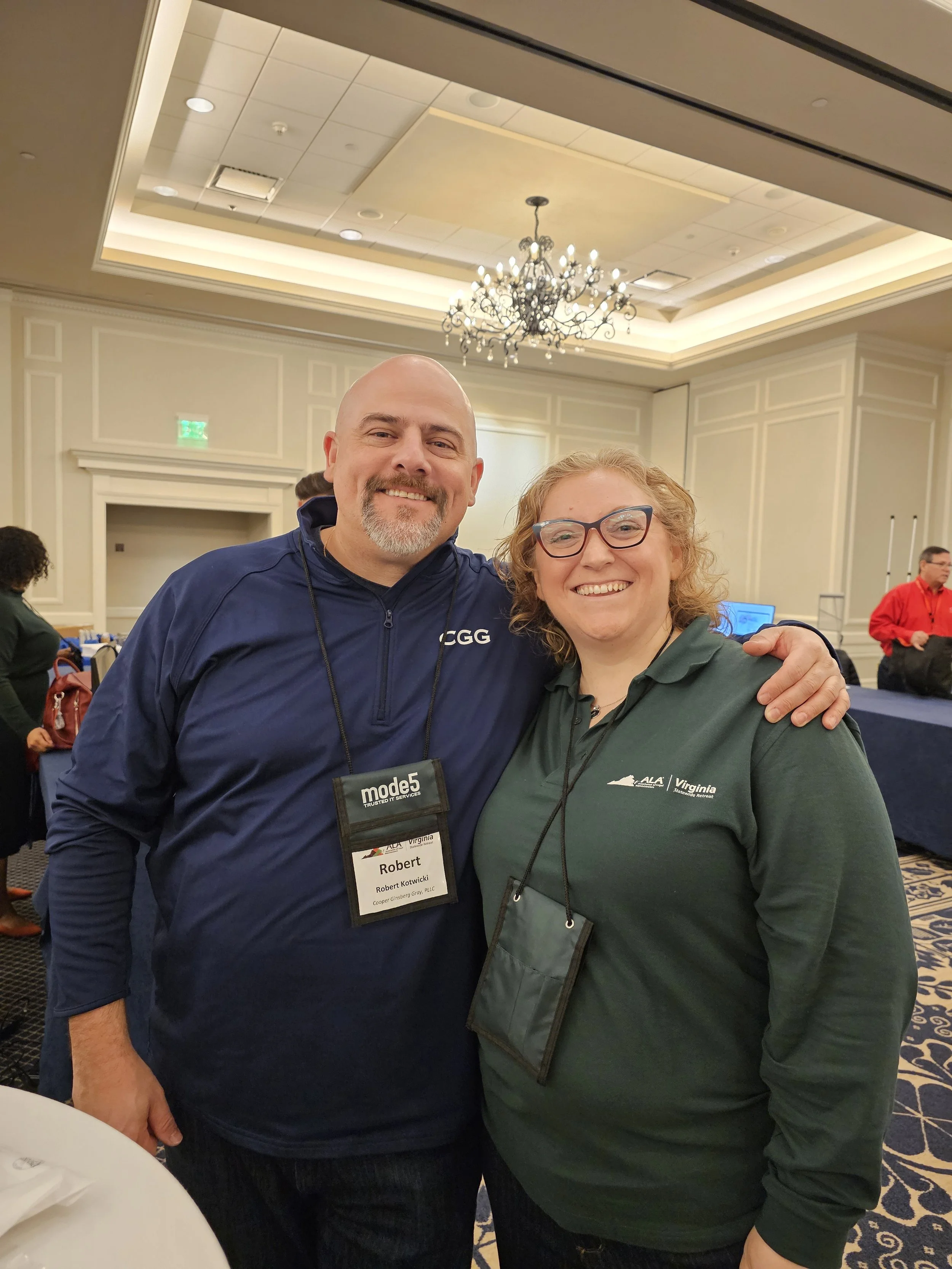 Two smiling people standing close together at an indoor event, with a chandelier and formal decor in the background. One is a man with a badge that reads 'Robert' and the other a woman with curly hair and glasses.