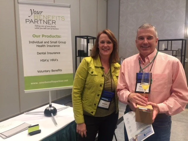 Two smiling people standing next to a health insurance booth at a conference. The woman is wearing a yellow jacket and the man is holding a glass of champagne and a small box.