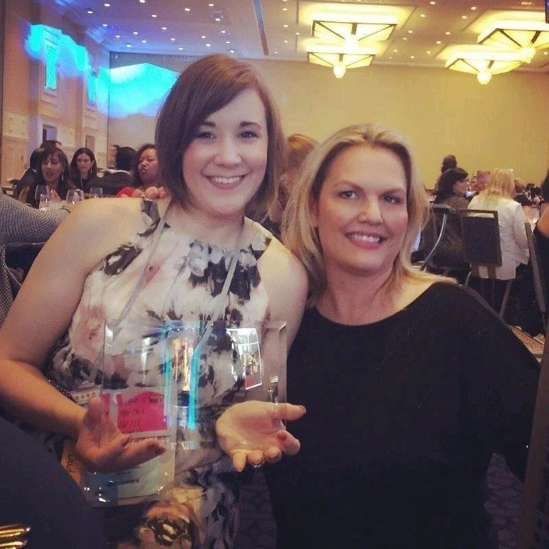 Two women at a formal event, smiling and holding a clear glass award.