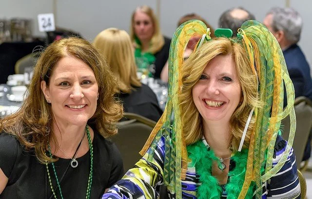 Two women smiling at an event, one wearing decorated green and yellow streamers on her head, with other people sitting at tables in the background.
