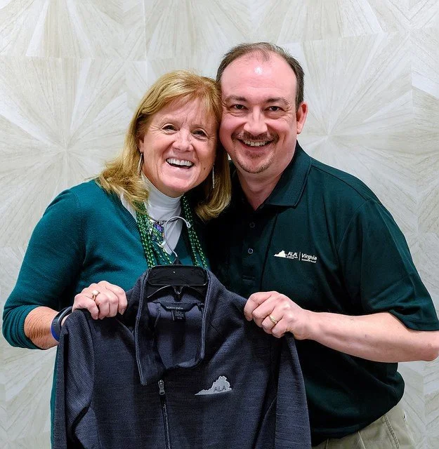 A smiling woman with light brown hair wearing a teal sweater and white turtleneck, and a man with short dark hair and a beard in a black polo shirt, holding a black fleece jacket with a mountain logo and the word "Virginia", standing against a light-