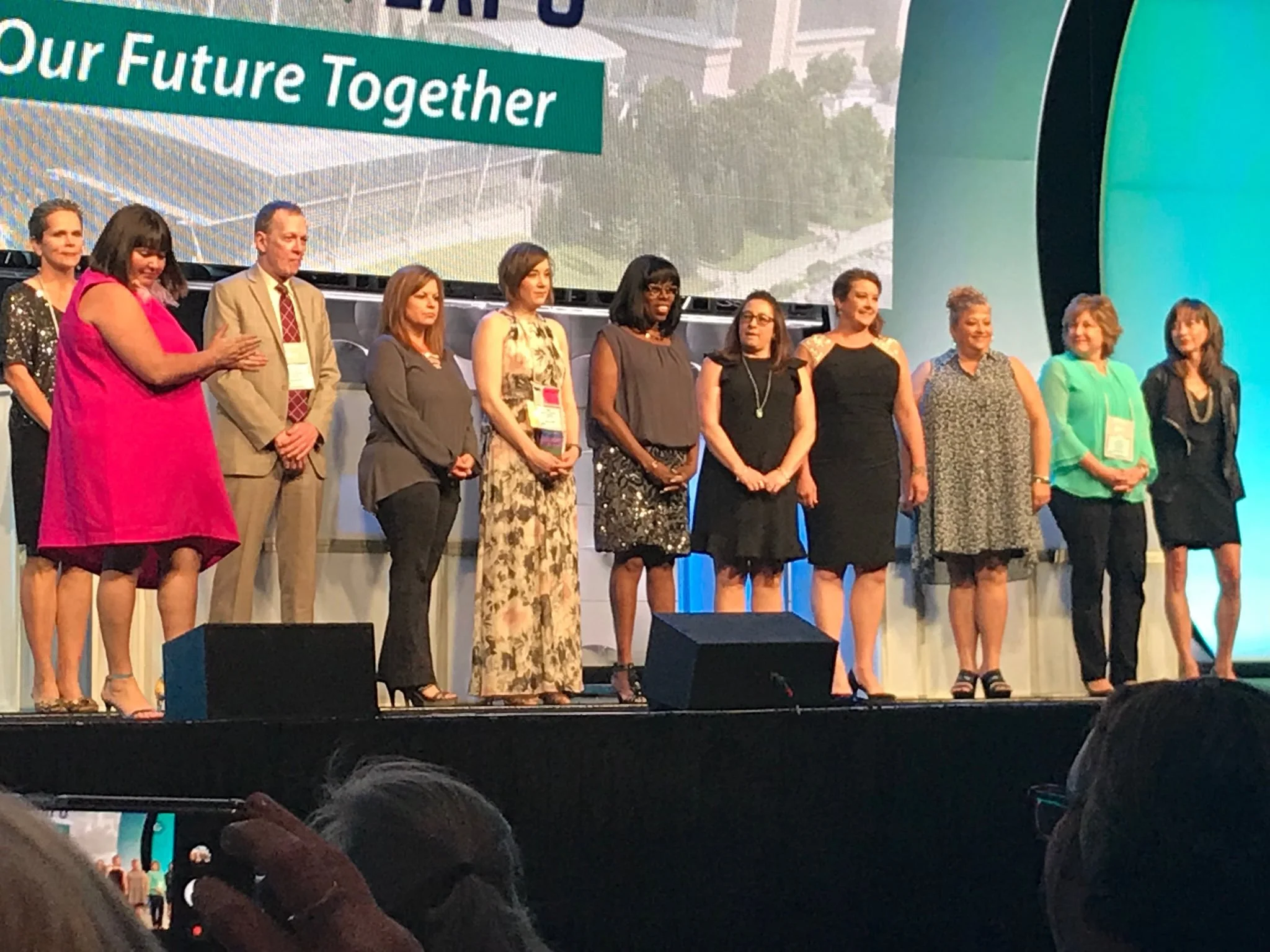 A group of people, mostly women, standing on a stage at a conference or event, with a large screen behind them displaying the text 'Our Future Together' and an image of a cityscape.