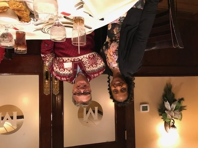Two women sitting at a dinner table with glasses and plates, smiling at the camera in a festive setting with holiday decorations.