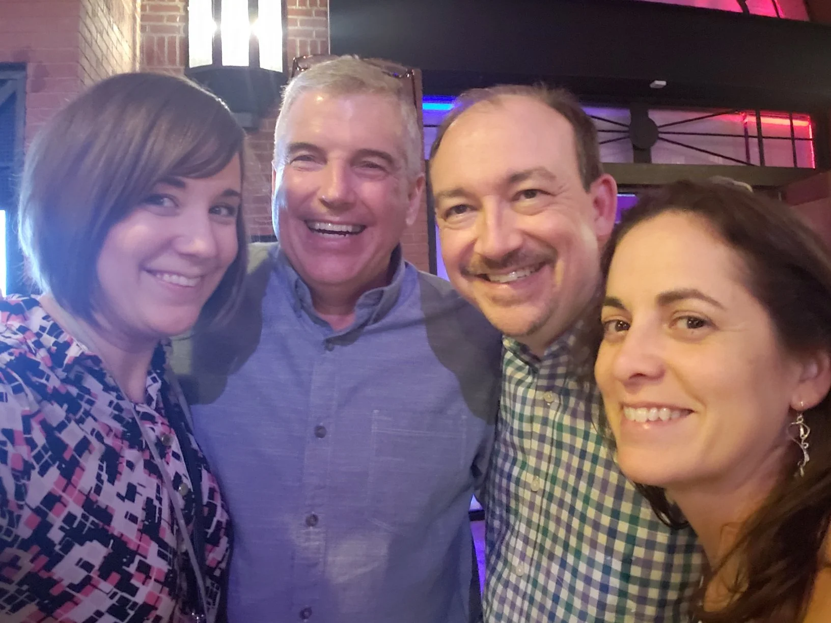 Group of four friends smiling and taking a selfie at a bar or restaurant with dim lighting and brick walls.