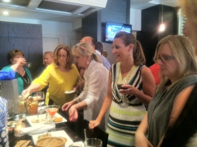 Group of women gathered around a bar, socializing, with drinks in hand in a lively indoor setting, possibly a restaurant or bar.