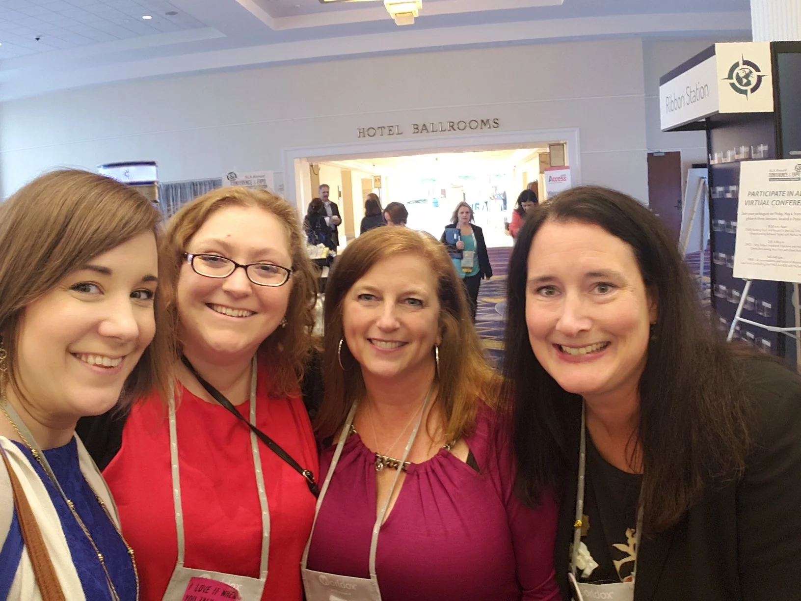 Four women smiling at the camera at a hotel conference lobby with signs for hotel ballrooms and ribbon station in the background.