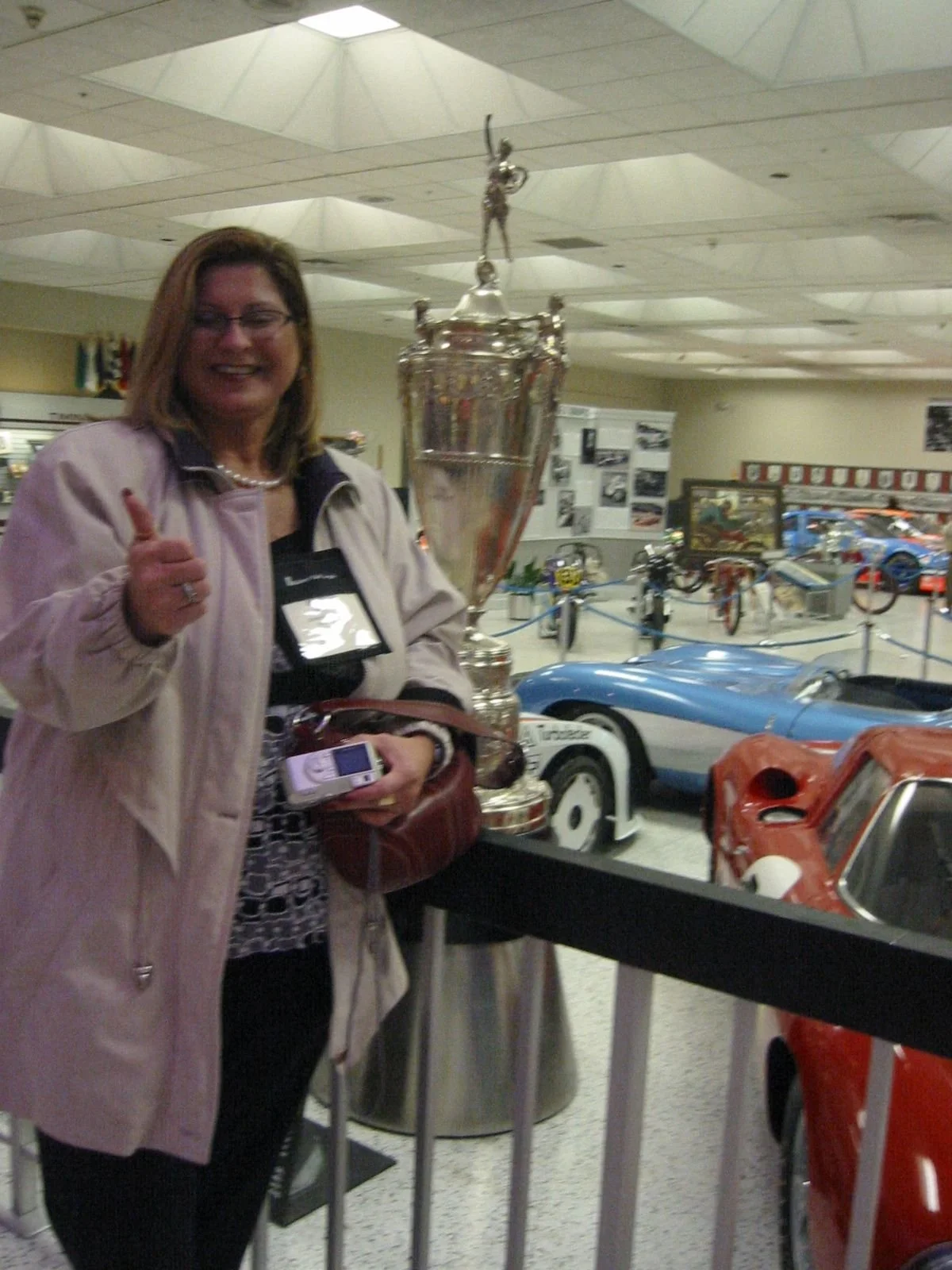 A woman smiling and giving a thumbs-up standing next to a large trophy inside a museum with vintage race cars in the background.
