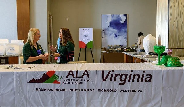 Two women talking behind a table with an ALA Virginia banner, with a buffet and a person in the background in a conference room.