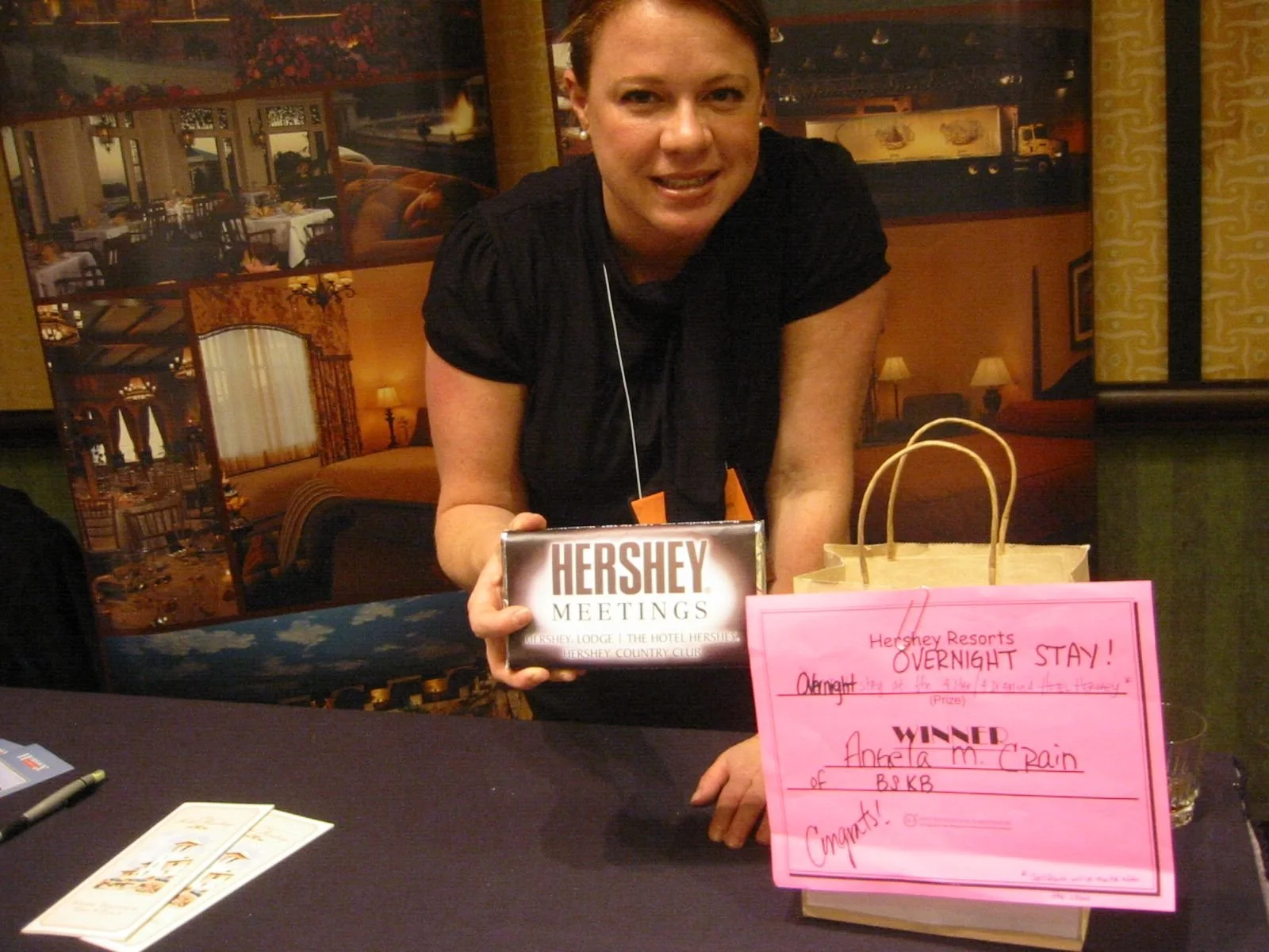 A woman at a Hershey's meeting holding a Hershey's chocolate bar, on a table with a pink sign that reads 'Overnight Stay Winner.' The background is decorated like a hotel with paintings of dining rooms and a bedroom.