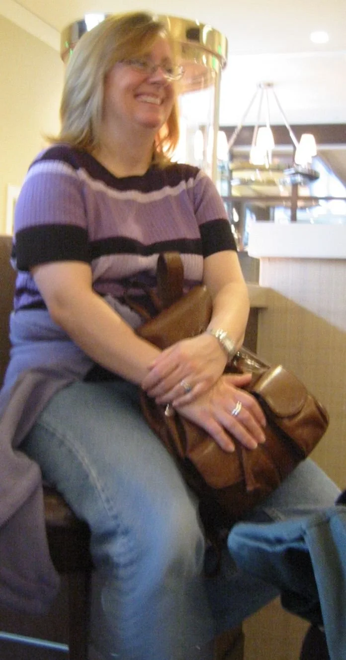 A woman with shoulder-length blonde hair, wearing glasses, a purple and black striped shirt, and light-colored jeans, sitting in a cafe or restaurant with a brown purse in her lap and smiling.