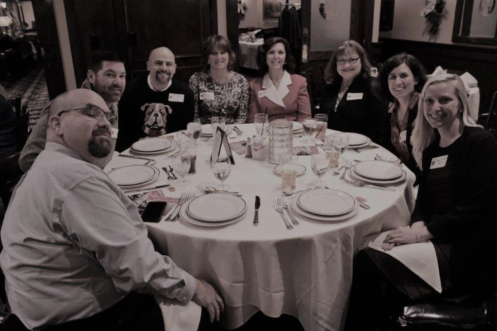 Group of nine people sitting around a round dining table at a restaurant, smiling and posing for the camera.