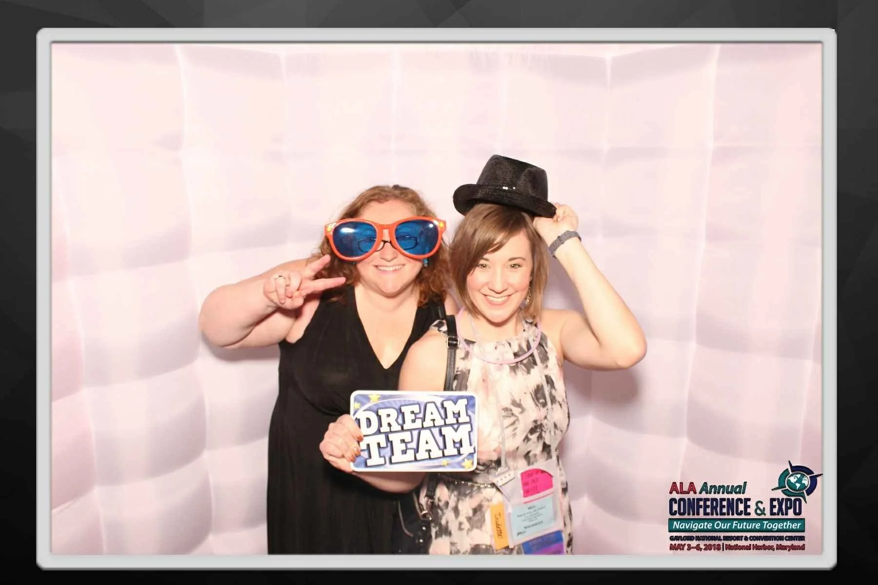 Two women smiling and posing with photo props at a conference, one wearing large blue sunglasses and making a peace sign, and the other wearing a black hat and holding a sign that says "Dream Team." The backdrop is a white, padded wall, and there is 