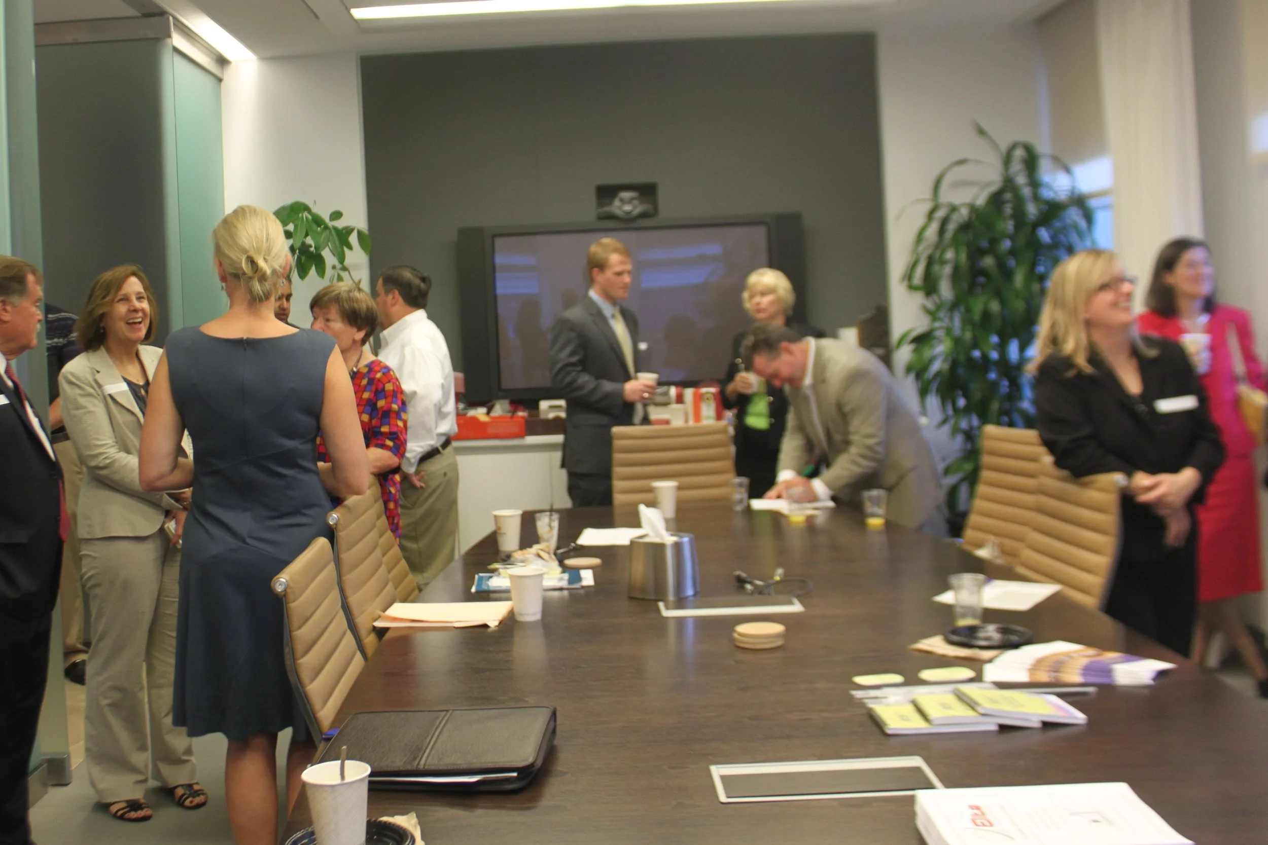Business meeting room with people socializing, standing around a large conference table with laptops, papers, and coffee cups, in an office setting.