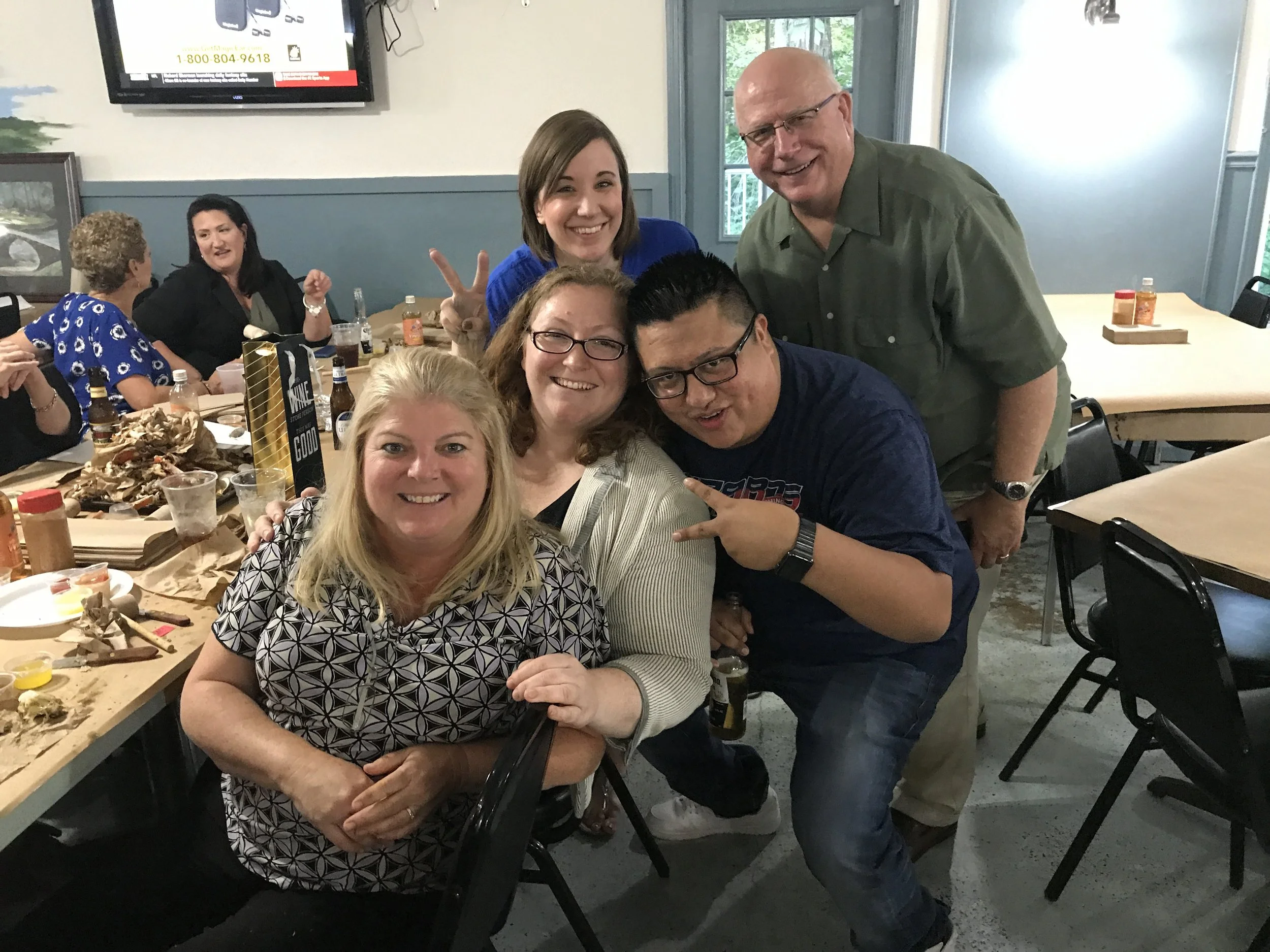 Group of people at a social gathering enjoying food, drinks, and conversation in a dining setting, smiling and posing for the camera.