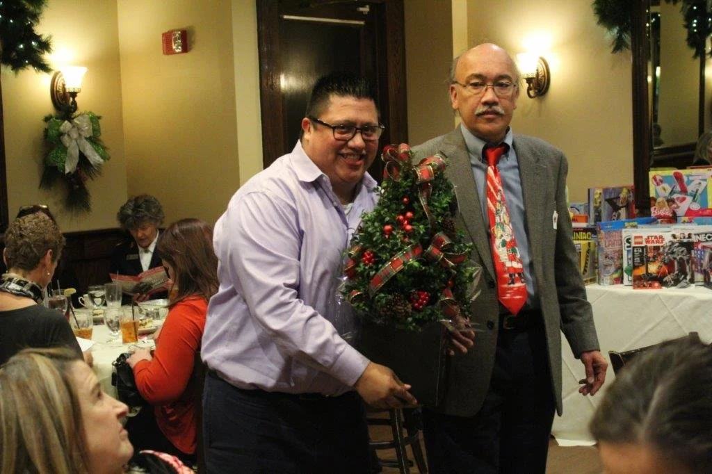 Two men at a holiday gathering, one holding a small decorated Christmas tree. Several people seated at a table in the background, some reading or drinking, in a warmly lit room decorated with Christmas wreaths.