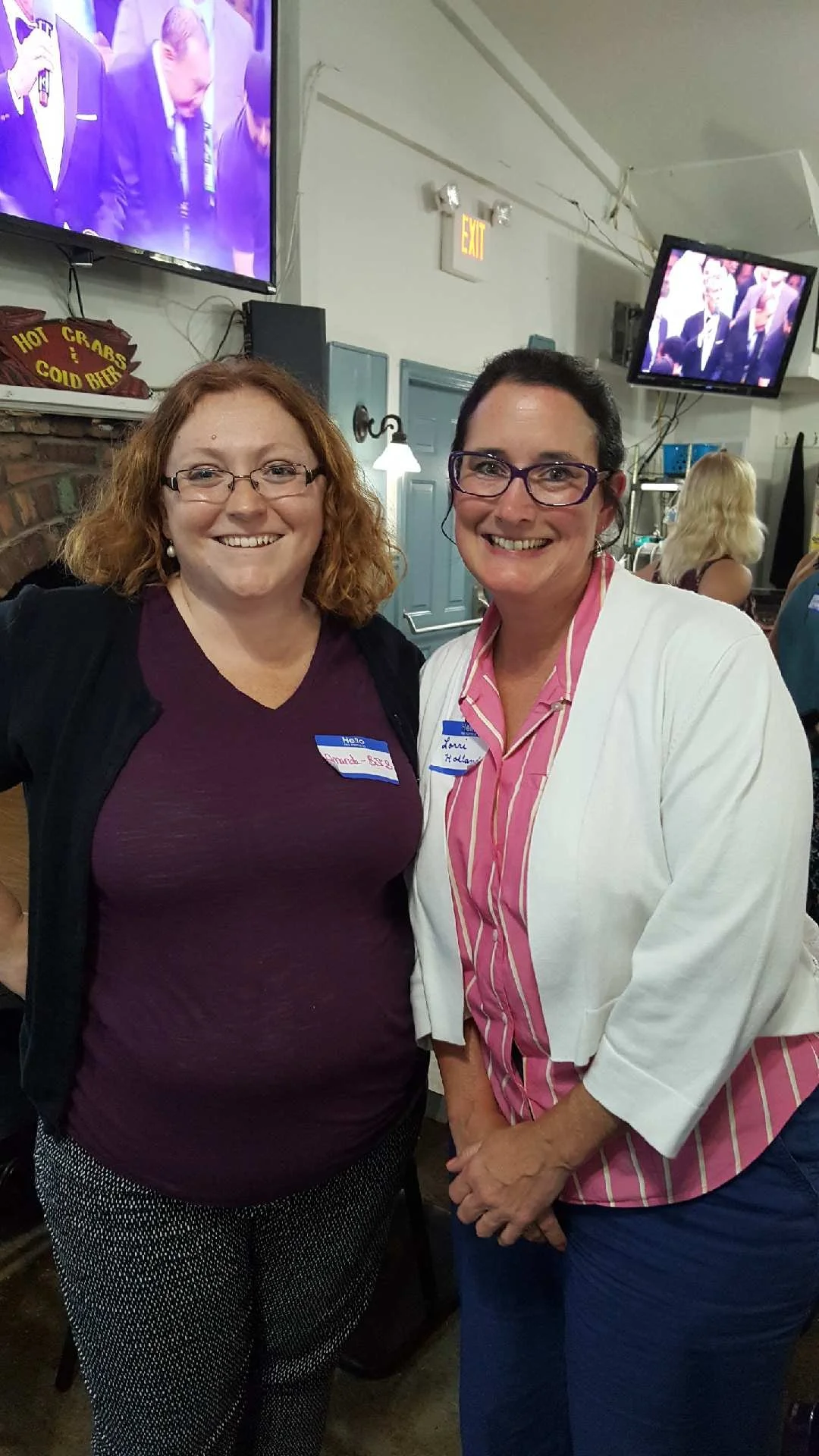 Two women smiling at a social gathering. One has red hair, glasses, and wearing a dark purple top with a name tag. The other has black hair, glasses, and wearing a pink and white striped blouse with a white blazer and a name tag.