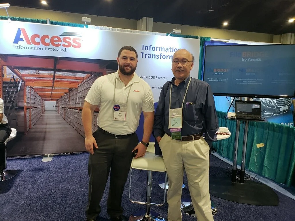 Two men standing in front of an exhibition booth with a large banner that reads "Access" and "Information Protected." The booth has a background of warehouse shelving with stored boxes and a display screen.