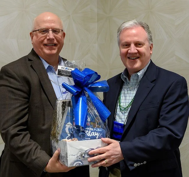 Two smiling men in suits holding a gift basket wrapped with a blue ribbon and bow, at an indoor event.