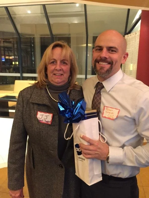 A woman and a man smiling, holding a gift bag with a blue ribbon, at an indoor event with glass windows behind them.