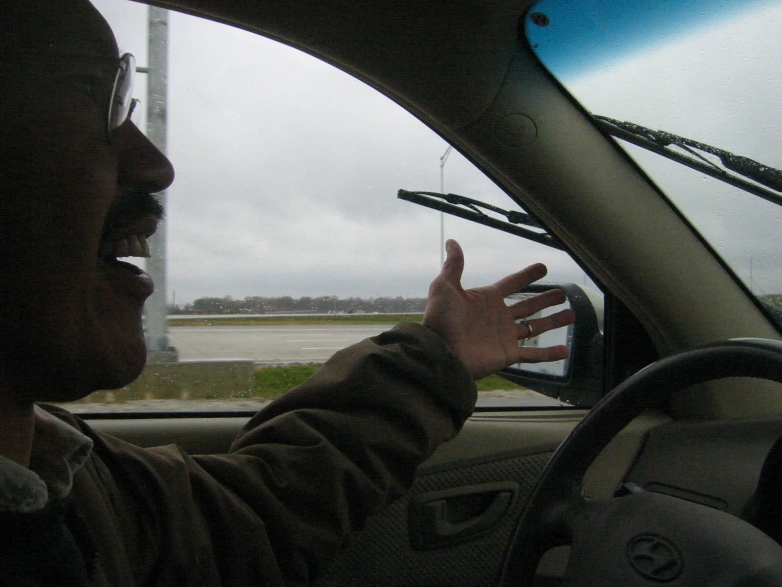 A person in the driver's seat of a vehicle, wearing glasses and a brown jacket, gesturing with their right hand while talking. Outside the window, overcast weather with rain and an open road are visible.