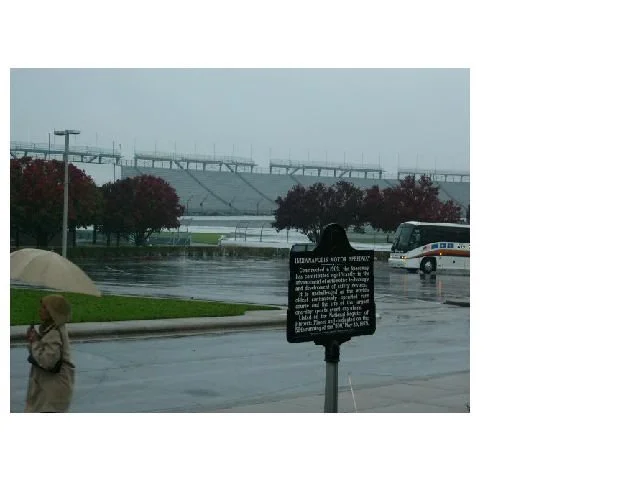 Rainy day scene outside a stadium with person holding umbrella, parked bus, trees with red leaves, and informational sign in foreground.