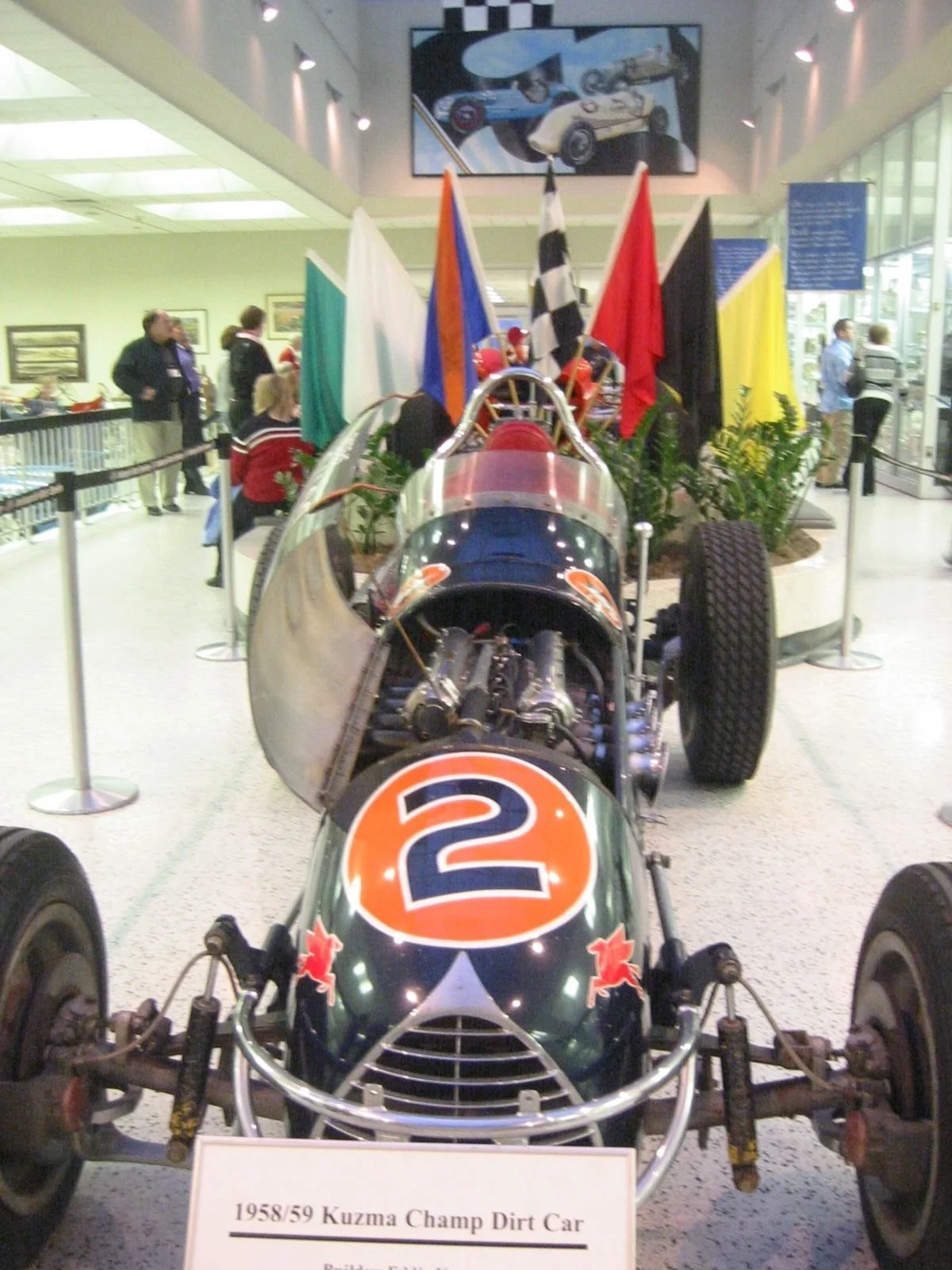 A 1958/59 Kuzma Champ Dirt Car race car on display at a museum, with flags and people in the background.