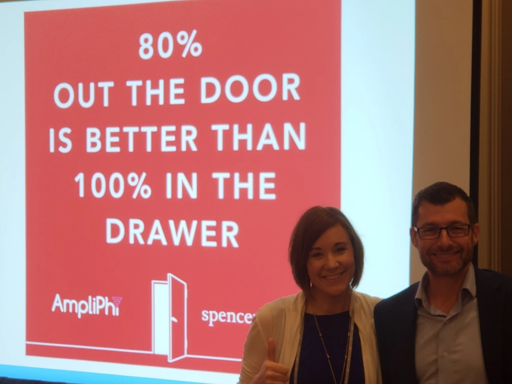 Two people, a woman with dark hair and a man with glasses, standing in front of a red and white presentation slide with text about '80% out the door is better than 100% in the drawer,' and company logos for Ampliphi and spencer.