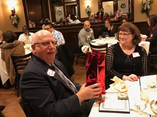 An elderly man in a suit holding a red gift bag at a dinner event, sitting next to a woman with glasses, in a warmly lit restaurant filled with other guests.