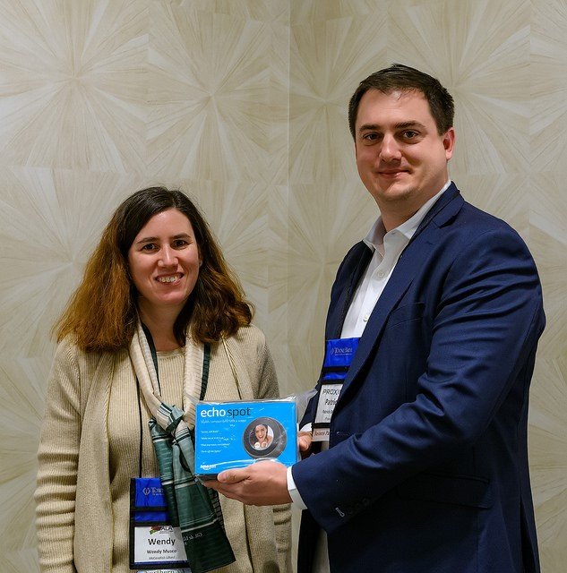 A man in a dark blue suit holding a blue box with a photo of a woman on it, standing next to a woman with brown hair wearing a beige coat and scarf, both smiling at the camera.