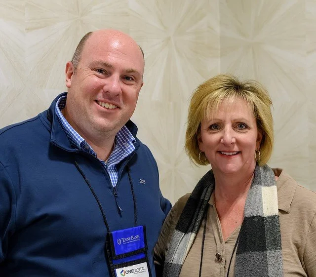 A man and a woman smiling, standing in front of a beige textured wall, dressed in casual professional attire, with the man wearing a blue zip-up jacket and the woman wearing a beige blazer and a gray scarf.