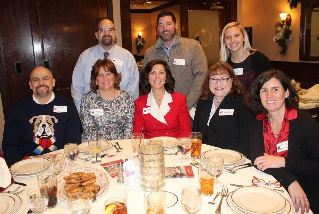 Group of nine people gathered around a dining table in a restaurant, smiling for the photo. Some have name tags, and the table has plates, glasses, and a tray of food.