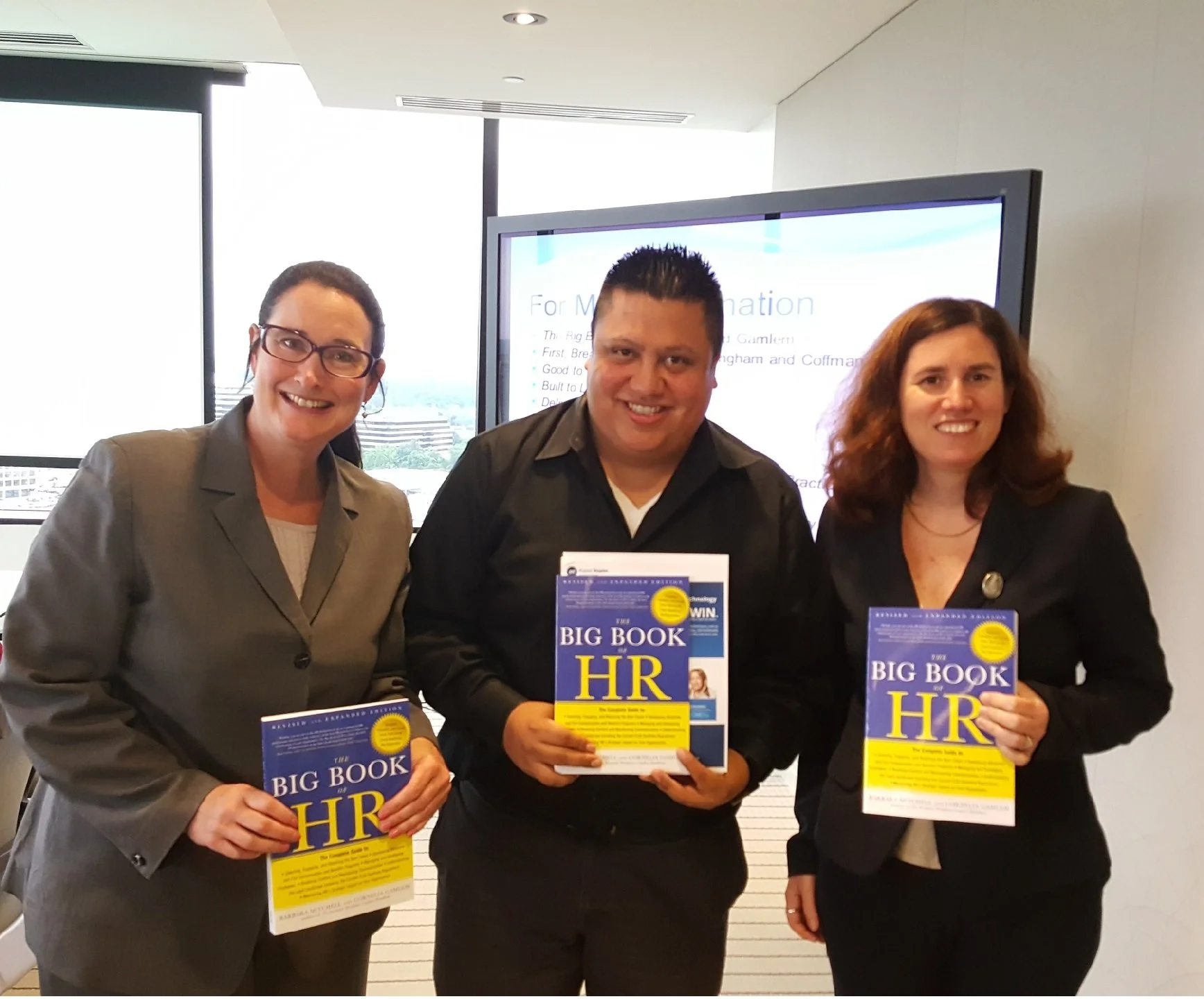 Three individuals smiling and holding copies of the book titled 'The Big Book of HR' at what appears to be a professional event or conference, standing in front of a large screen displaying presentation content.