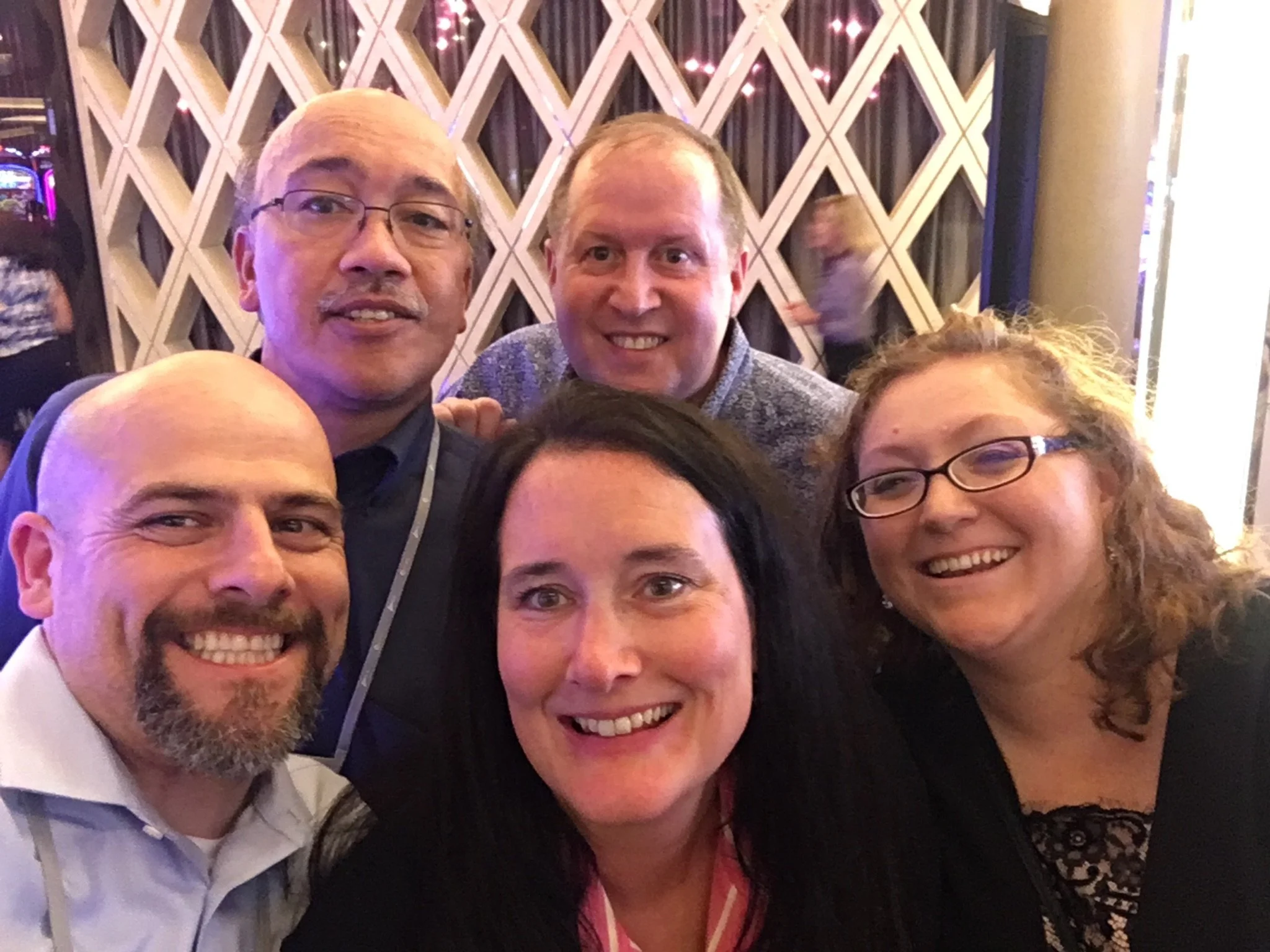 Group of five smiling people taking a selfie in front of a decorative background with geometric patterns and pink lights.