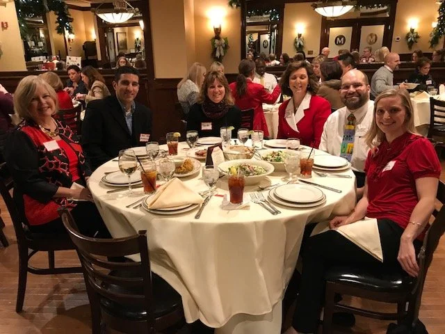 Group of six people sitting around a round table at a restaurant, with food and drinks, during a festive gathering.