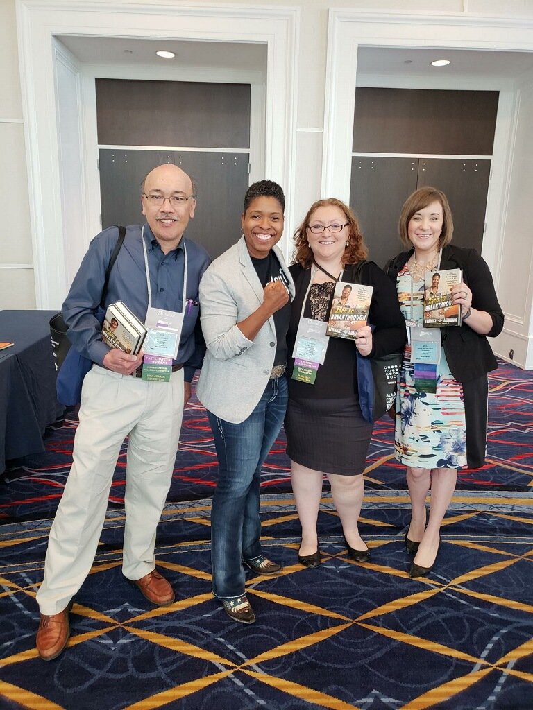 Four smiling women at a conference, holding books and wearing conference badges, standing on a patterned carpet in a large room with cream-colored walls.