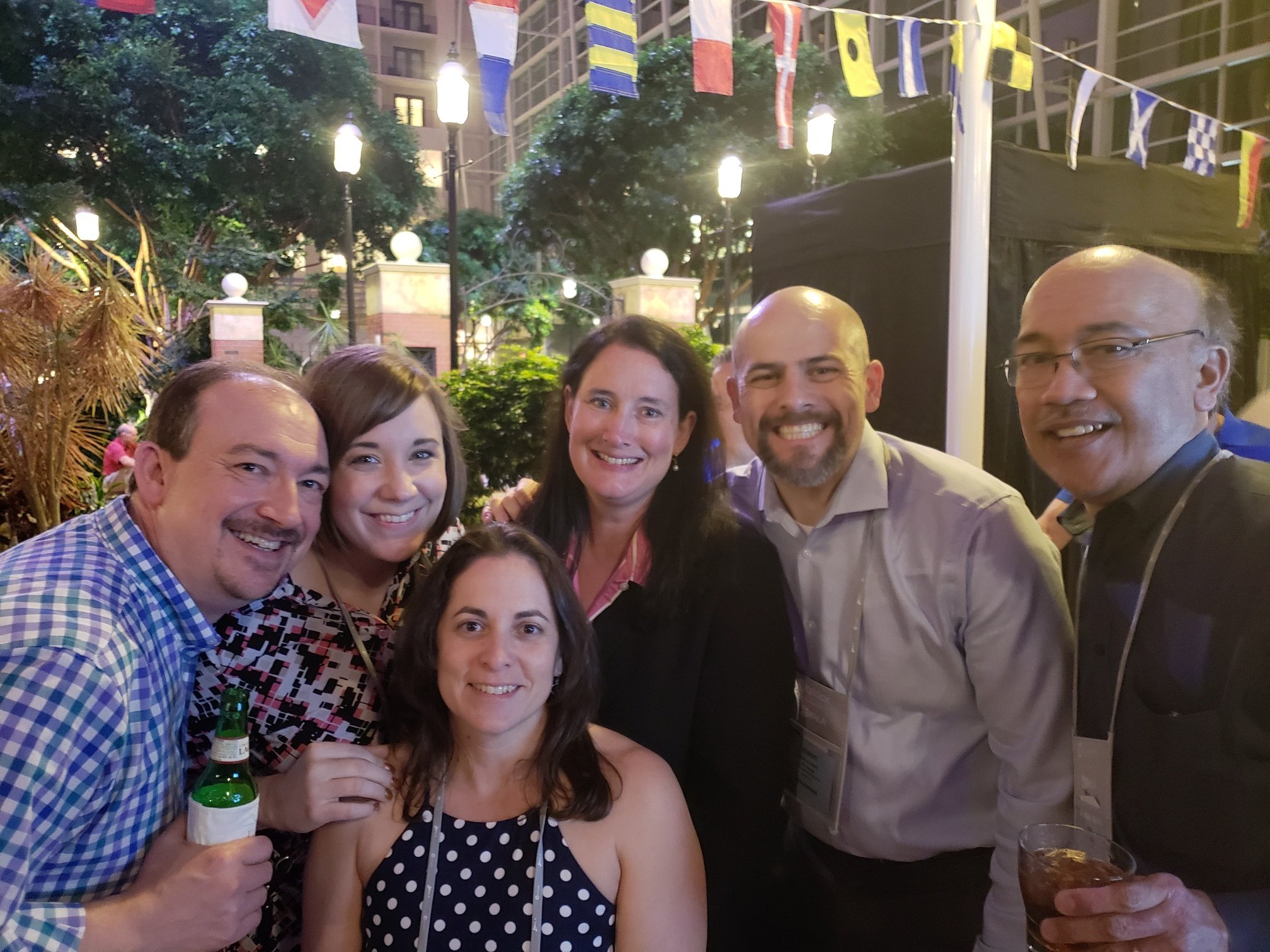 Group of five adults smiling at a social event outdoors at night, with string flags and decorative lights above, and a few trees and building in the background.