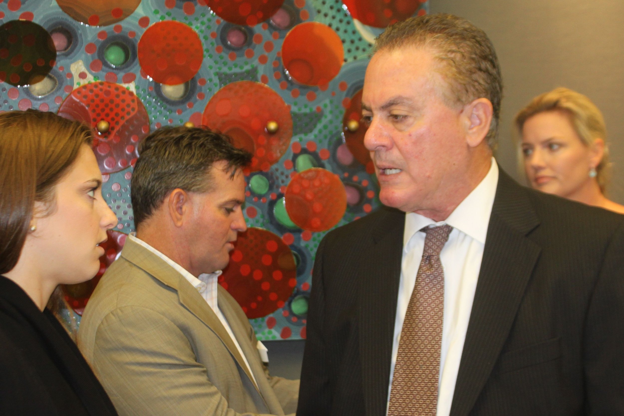 Four people, two men and two women, standing close in conversation indoors, with a colorful, abstract, red and green dotted artwork in the background.