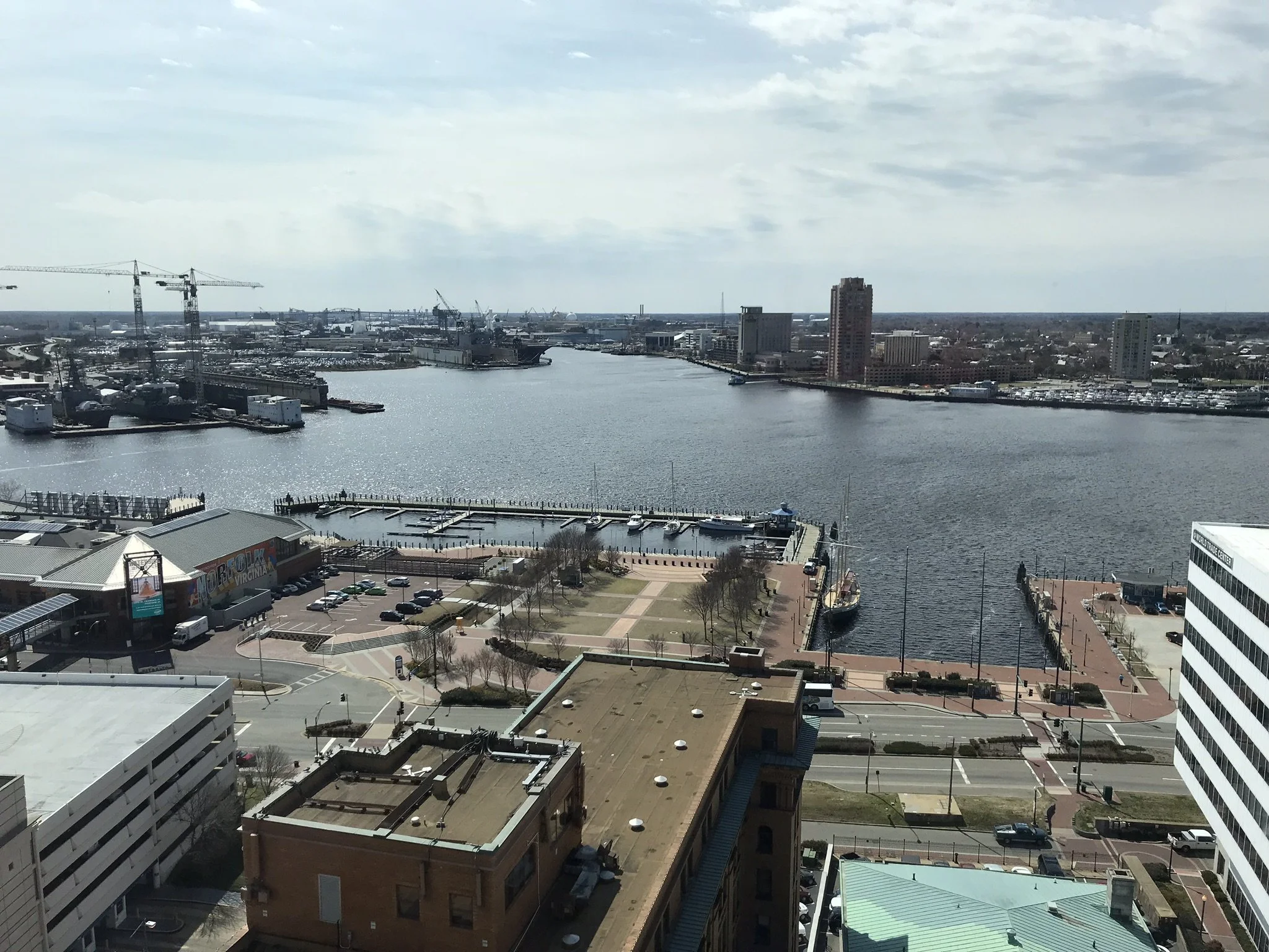 A cityscape with a river running through it, surrounded by tall buildings, construction cranes, and boat docks, under a partly cloudy sky.