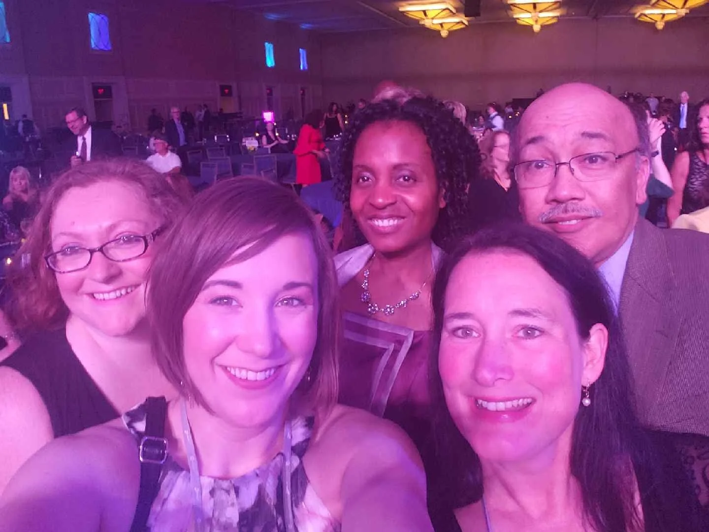 Group of five women and one man taking a selfie at a large indoor event with people in the background.