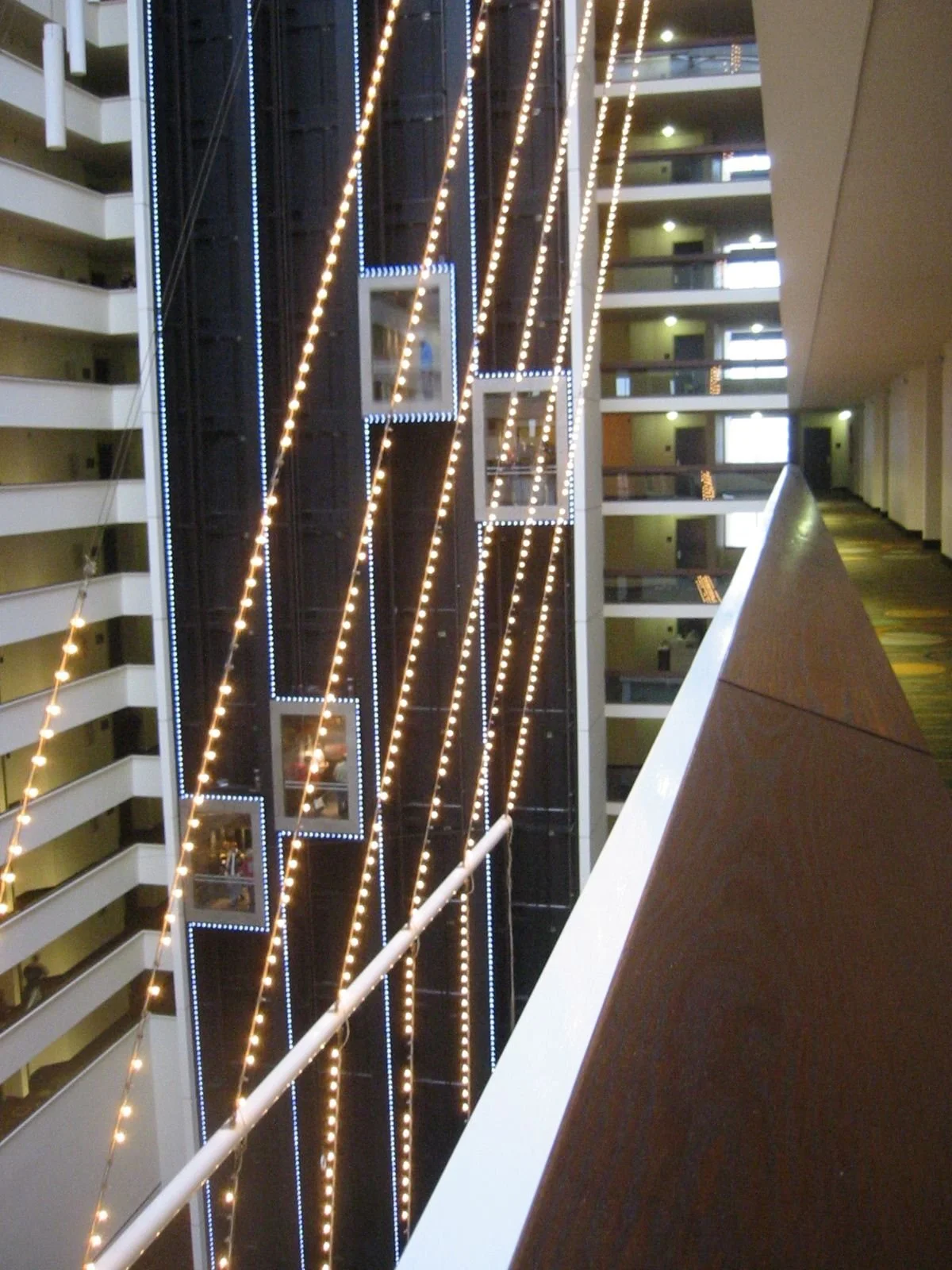 Interior view of a multi-floor hotel or apartment building with illuminated vertical light strips and several open elevator shafts, seen from a balcony.