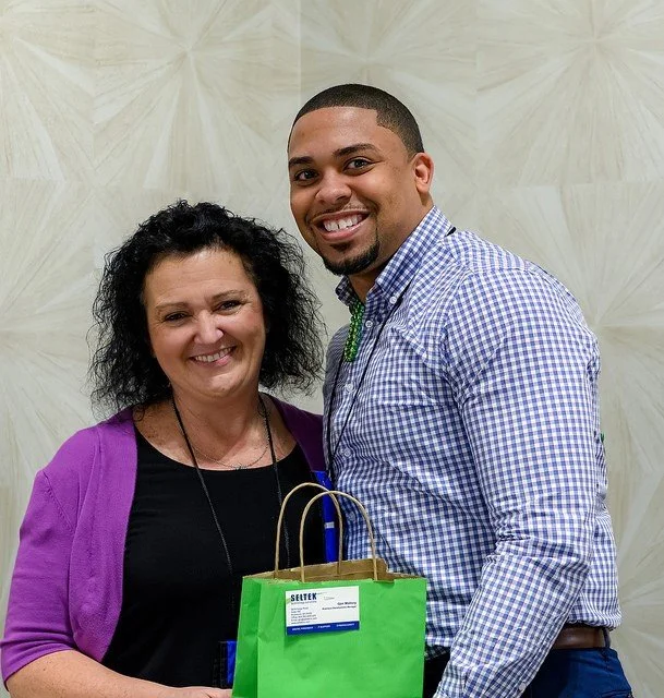 Two smiling people, a woman and a man, standing together with a green gift bag in front of them, indoors against a textured beige background.