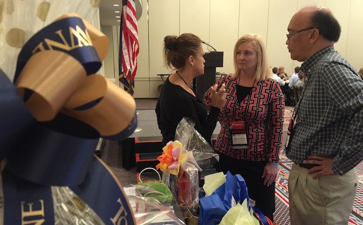 Three people engaged in a conversation at an indoor event, with gift bags and decorative items on a table, and an American flag in the background.