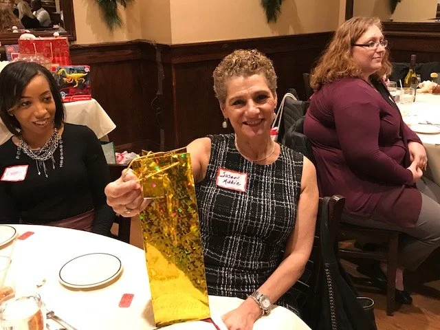 A woman smiling and holding a large yellow gift bag at a banquet table with other women seated nearby.